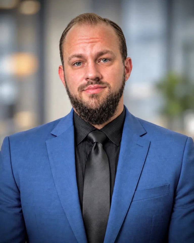 Professional headshot of a bearded man wearing a blue blazer and black dress shirt against a blurred office background